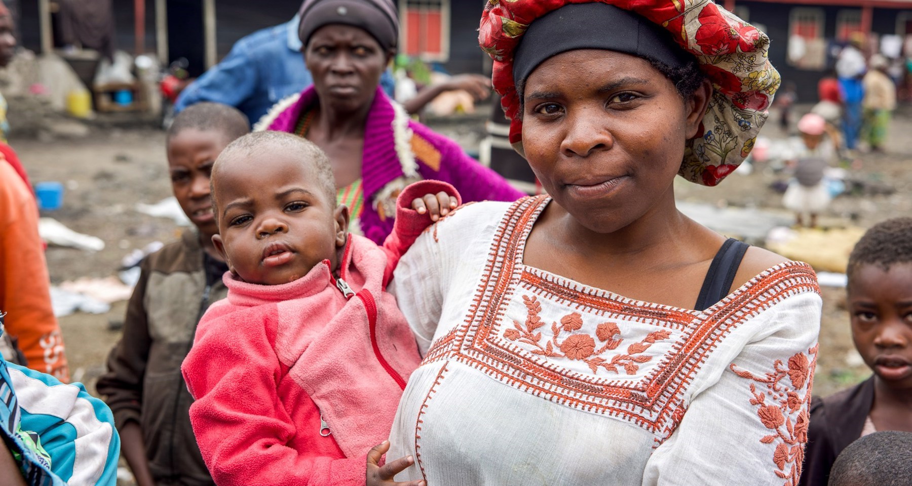 En kvinne med hodeplagg holder et barn på armen. I bakgrunnen ser vi andre mennesker og en skole i DR Kongo. 