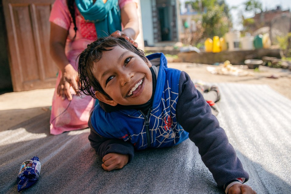 En smilende jente fra Nepal ligger på en matte på bakken. Bak henne sitter en kvinne med hånden kjærlig på hodet til jenta.