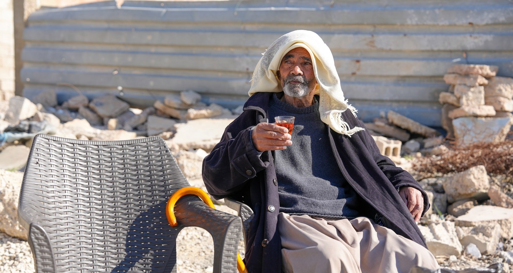 Eldre mann sitter på byggeplass med en kopp te i hånda og tørkle over hodet. 