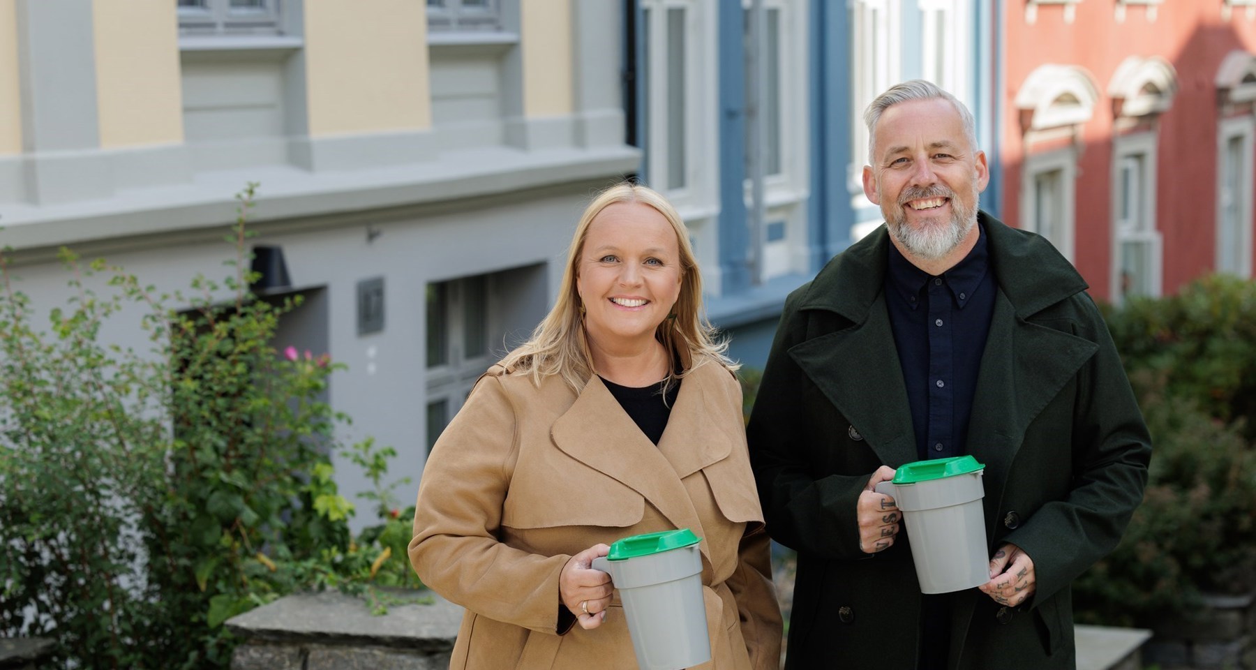 En blond kvinne i beige kåpe og en mann med skjegg og svart frakk står med bøsser utenfor hus i en by.