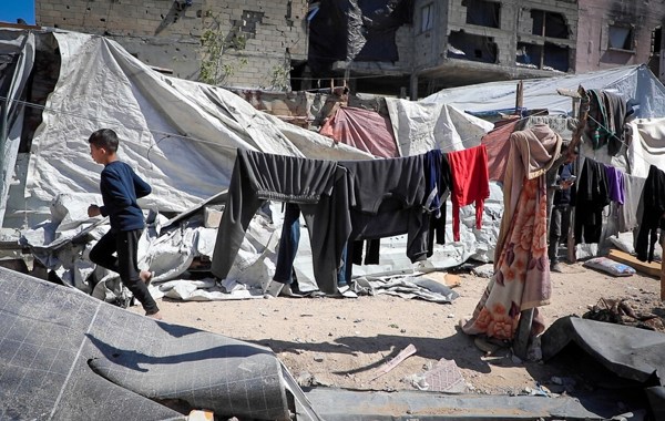 En ung gutt løper på en grusvei omringet av telt og klær på tørkesnorer. I bakgrunnen er det et murhus med store skader. 
