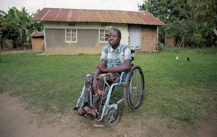 En ung, smilende gutt sitter i rullestol på gressplen med hus i bakgrunnen i Uganda. 