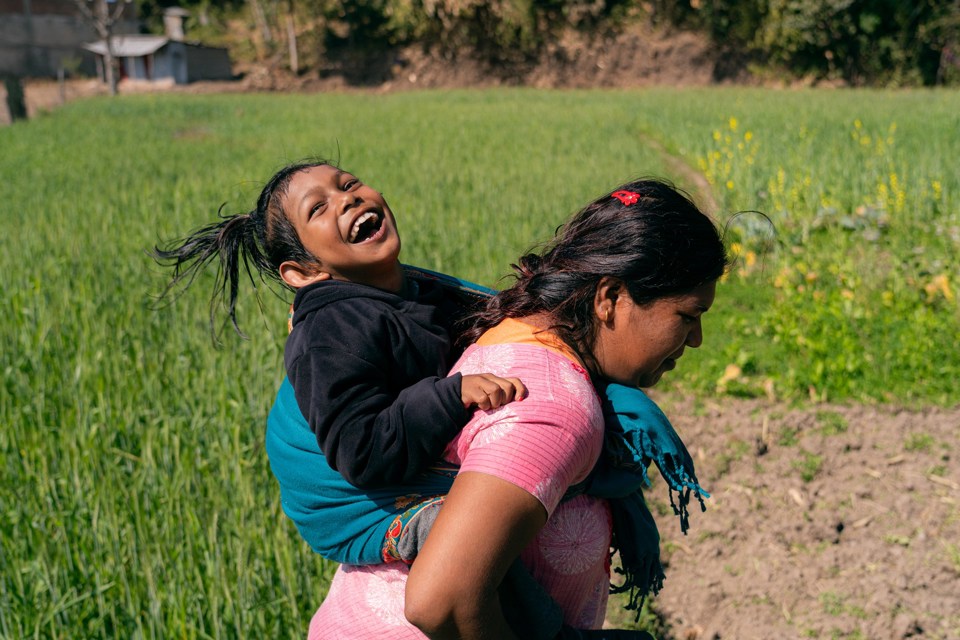 Jente sitter på ryggen til moren sin i en grønn åker i Nepal. Jenta smiler og ler.