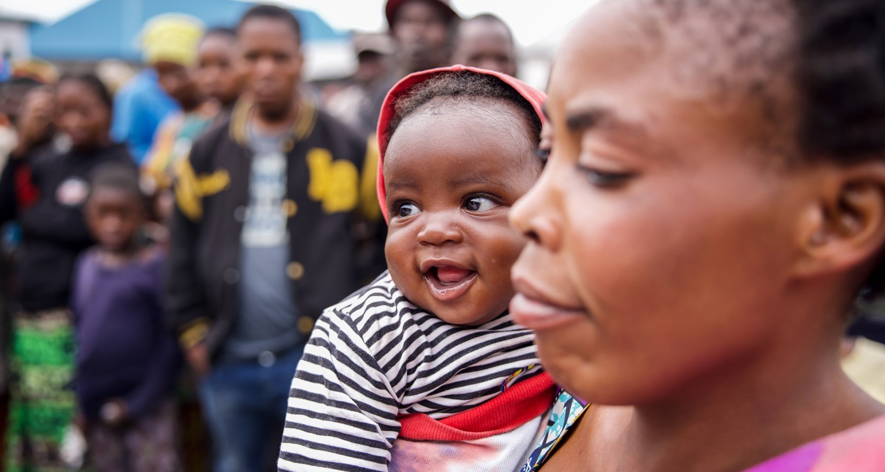En mor holder et barn som smiler på armen. I bakgrunnen ser vi flere mennesker og et slags telt. 