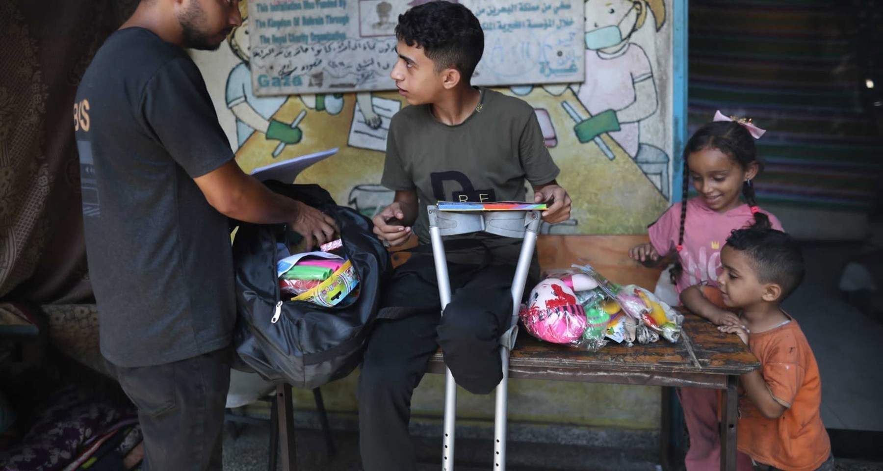 En gutt med amputert bein sitter på en benk. En voksen mann og to små barn står ved siden av.