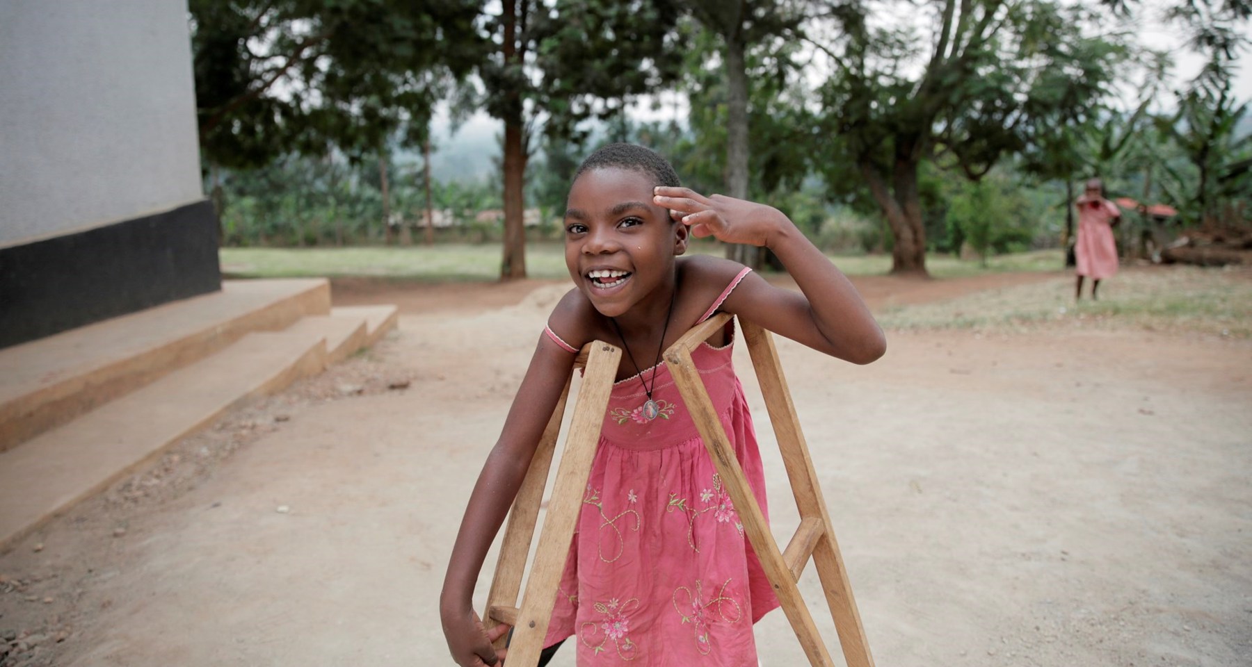 En smilende jente med rosa kjole står ute med krykker og løftet hånd.