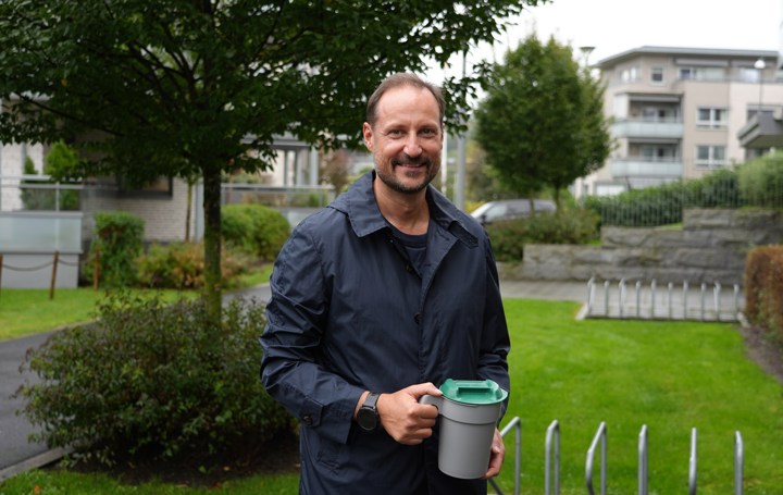 Kronprins Haakon står smilende med blå jakke i et boligområde med en bøsse i hendene. 